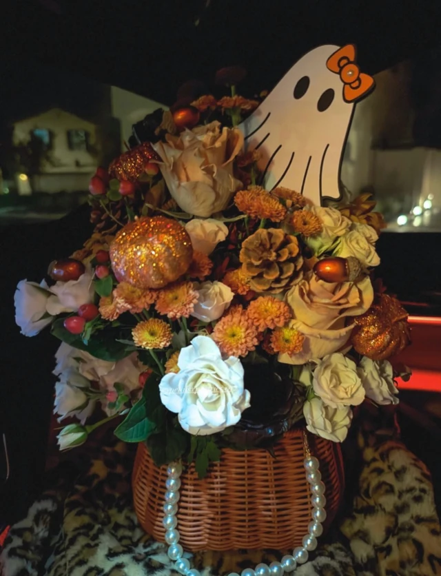 A woven basket of cream roses and orange flowers with a ghost decoration and glitter pumpkins