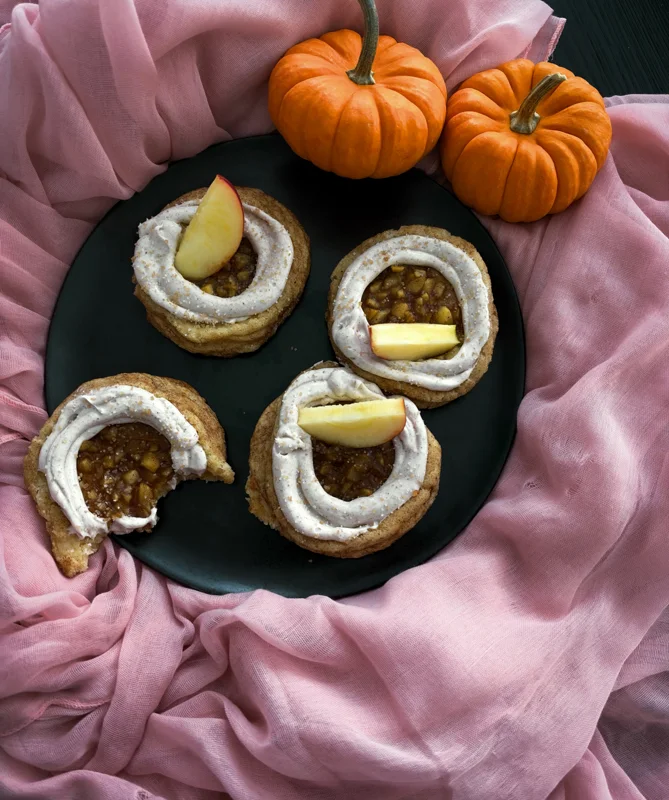 Autumn treats topped with cream and apple slices on a black platter beside small pumpkins